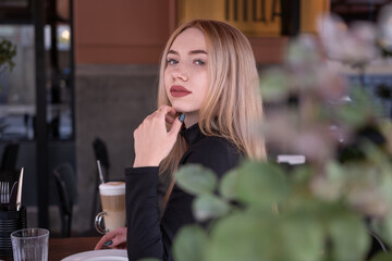 Pensive attractive young blond woman sits at table in cafe and looks into camera. Portrait of young girl in summer cafe