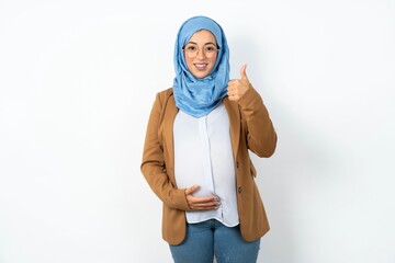 Young beautiful pregnant muslim woman wearing hijab in white studio doing happy thumbs up gesture with hand. Approving expression looking at the camera showing success.