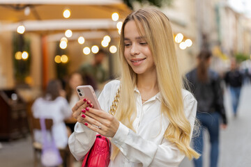Tourist traveler blonde young woman using smartphone typing text messages looking for search a way on map in mobile navigator app outdoors. Teenager girl walking passes by urban city street background