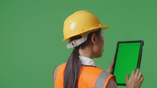 Close Up Back View Of Asian Female Engineer With Safety Helmet Working On A Green Screen Tablet And Looking Around While Standing In The Green Screen Background Studio
