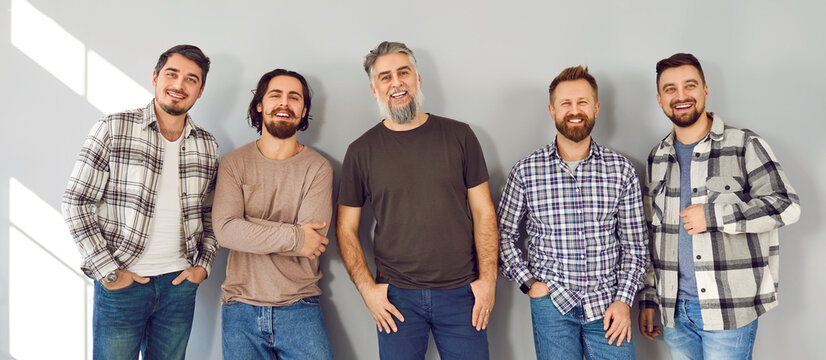 Happy Men In Casual Clothes. Group Of Cheerful Male Fashion Models Of Different Ages Wearing Quiet Subdued Color Plain Tshirts Or Plaid Shirts Standing By Gray Background Looking At Camera And Smiling