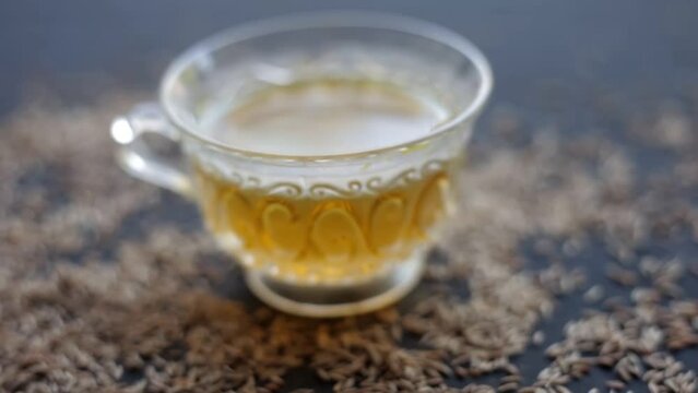 Hd footage 24 FPS. Cinematic close-up shot of cumin seeds or zeera aka jeera (Cuminum cyminum) in a glass bowl along with its herbal tea in a cup next to it on glossy surface, 