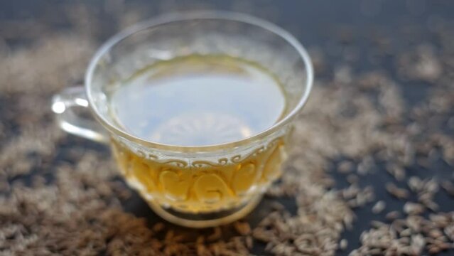 Hd footage 24 FPS. Cinematic close-up shot of cumin seeds or zeera aka jeera (Cuminum cyminum) in a glass bowl along with its herbal tea in a cup next to it on glossy surface, 