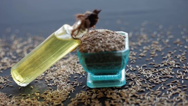 HD shot of jeera oil in a transparent bottle along with raw cumin seeds (Cuminum cyminum) on a black wooden surface.
