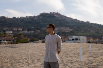 young caucasian man in a collarless shirt and dress pants, on the beach looking to the side