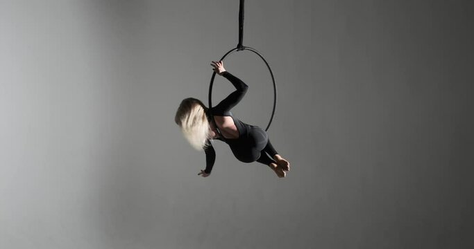 A caucasian woman, dressed in a form-fitting black jumpsuit on an aerial hoop against a dark background. With long hair, she combination of strength, agility as she moves through the air.