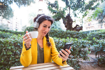 Woman with headphones looks at her phone while holding a cup of coffee on a park terrace
