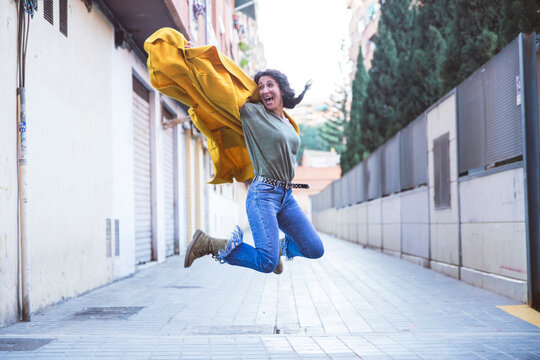 Adult Woman In A Yellow Coat Jumps On An Urban Street