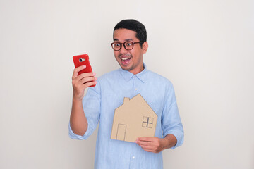 A man with wow expression looking to mobile phone and holding house shape cardboard - obrazy, fototapety, plakaty