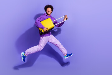 Full size photo of funny funky handsome person dressed violet hoodie pants hold valise run to airport isolated on purple color background