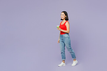 Full body young woman of Asian ethnicity wearing casual clothes red tank shirt hold takeaway delivery craft paper brown cup coffee to go walk isolated on plain pastel light purple background studio.