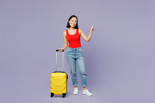 Traveler happy woman wear red casual clothes hold suitcase point aside isolated on plain pastel purple background Tourist travel abroad in free spare time rest getaway Air flight trip journey concept