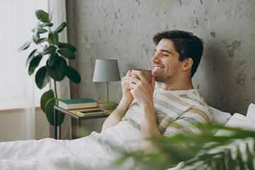 Profile happy young man wear casual clothes t-shirt pajama lying in bed drink coffee tea hold cup...