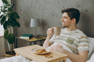Side view young man in casual clothes t-shirt pajama lying in bed eat cookies drink coffee tea have breakfast rest relax spend time in bedroom home in own room hotel wake up dream be in good mood day.