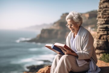 Medium shot portrait photography of a satisfied old woman reading a book against a scenic ocean cliff background. With generative AI technology