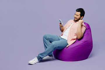 Full body fun young Indian man he wear pink shirt white t-shirt casual clothes sit in bag chair do winner gesture isolated on plain pastel light purple background studio portrait. Lifestyle concept.