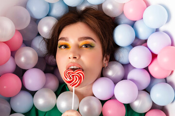 Fototapeta na wymiar portrait face shot, attractive beautiful girl stylish with bright makeup and double bun hair, emotional, facial expression, lying in a pool filled with pastel multi colored plastic balls, top view.