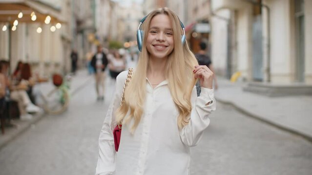 Rear view of blonde young woman tourist walking through the European street outdoors, turning to camera and smiling. Back view of teenager girl traveler walk in urban city background. Town lifestyles