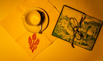 romantic candlelight scene of a white porcelain tea set, an old book open to an illustration, glasses, all resting on a light-colored linen placemat embroidered with a red ear of corn