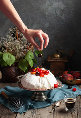 a woman's hand puts a cherry on a meringue homemade cake with sweet cherries and chocolate syrup on a dark background surrounded by vintage crockery and flowers - 611049154