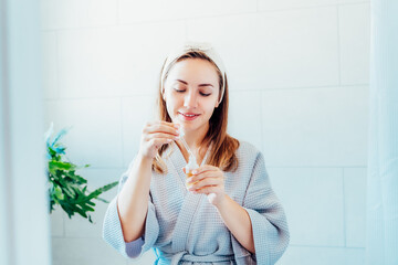 Obraz na płótnie Canvas Young woman in bathrobe looking in the mirror, holding dropper with hyaluronic acid serum close to face and smiling. Skin hydrating. Cosmetic spa procedures. Beauty self-care at home. Selective focus