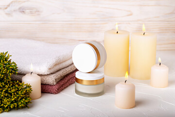 Fototapeta na wymiar Jars with day and night face cream, burning candles, towels and juniper branch on light background. Cosmetic skin care, spa procedures. Concept of calmness, comfort. Selective focus