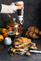 A female hand pours caramel syrup over a chocolate croissant spread with peanut butter and decorated with blueberries on a dark background. Breakfast concept. - 611050306