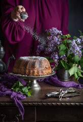 A woman's hand sprinkles powdered sugar on a homemade bundt cake on a table decorated with lilacs and vintage cutlery - 611050961