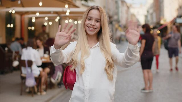 Blonde young woman smiling friendly at camera, waving hands gesturing hello, hi, greeting or goodbye, welcoming with hospitable expression outdoors. Teenager girl walking in urban city town street