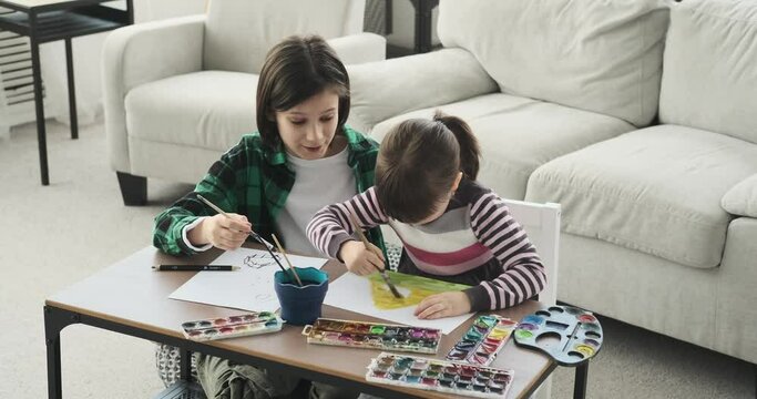 A pair of Caucasian siblings seated on the floor beside a small table at living room, they dive into the realm of art, armed with pencils and watercolors.