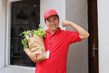 Happy excited male courier standing with a package of fresh food showing biceps muscle. Express...