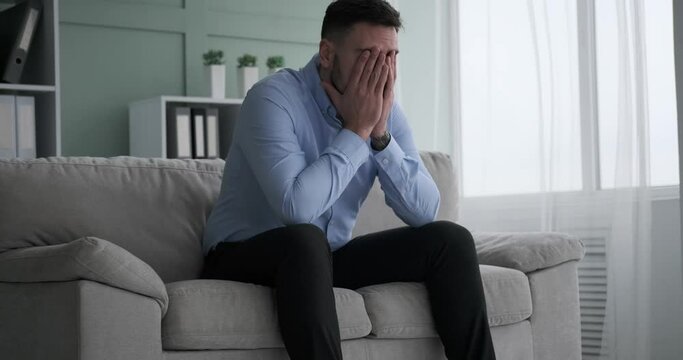 An exhausted Caucasian office worker of middle eastern descent comes home, collapses on the couch and holds his head due to a throbbing headache caused by overworking and stress.