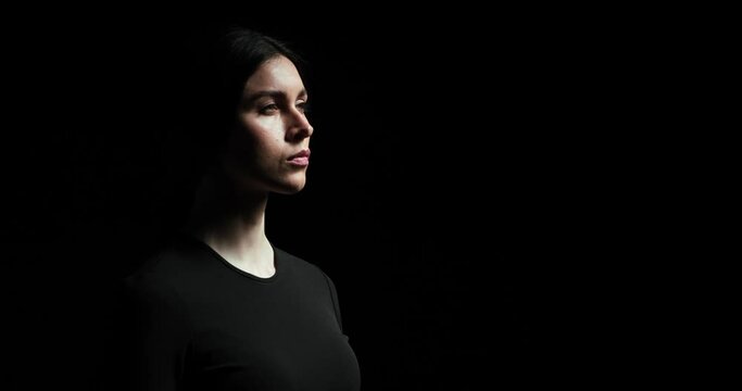 Serious and confident Caucasian woman wearing blouse is captured in profile on a black background. She lifts her head up and gazes towards the light with a thoughtful expression.
