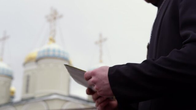 Person gives white post envelope to priest near church crop body on blurred background. Priest wears long black cassock with cross