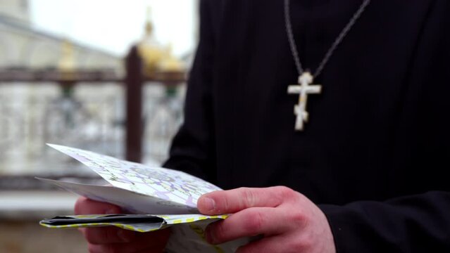 Priest stands and reads title of received card with invitation near church crop body on blurred background. Golden cross hangs on man cassock