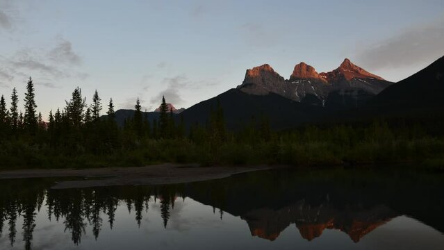 Timelapse sunset video of Three Sisters view seen in Canmore outside of Banff National Park with stunning reflection in water below. Clouds moving across sky with colours, blue sky, mountain peaks.