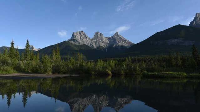 Timelapse sunset video of Three Sisters view seen in Canmore outside of Banff National Park with stunning reflection in water below. Clouds moving across sky with colours, blue sky, mountain peaks.