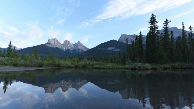 Panoramic sunset views over Three Sisters in Canmore, Alberta near Banff, Jasper National Park during summer time with afternoon, golden hour glow reflection in calm, pristine water below wilderness 