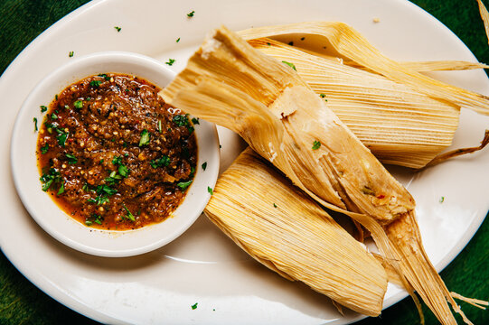 A platter of cooked wrapped tamales and a bowl of peppery salsa on a green table, closeup overhead