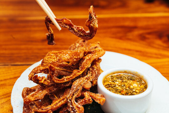 A pair of chopsticks hold crispy fried pigs ear above a plate of more with a spicy sauce on a white plate against a wooden surface