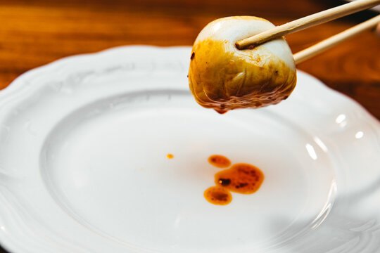 Pork bao dumpling held by chopsticks dripping sauce over an empty white plate against a wooden background