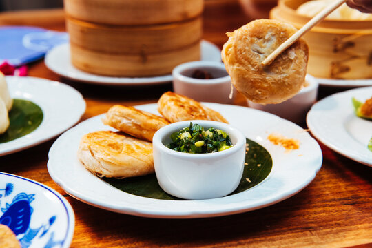 A pair of chopsticks holds a Chinese meat pie above a plate with more pies and scallions surrounded by other dim sum plates.