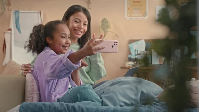 Happy 10 year old African American girl taking selfie portrait on smartphone with mom sitting together on bed in cozy kids bedroom with peach wallsHappy 10 year old African American girl taking selfie