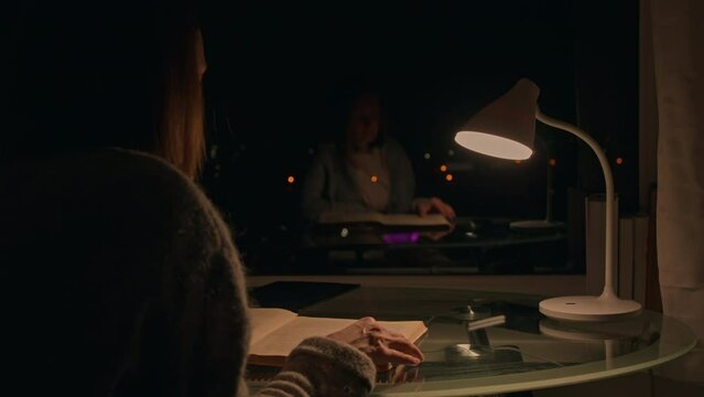 unrecognizable woman sitting at the desk with lamp reading book at night in apartment lady reflection in the window