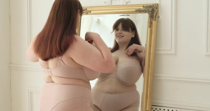 A plus-size Caucasian woman stands by the mirror in her bedroom, beaming with a smile. In this uplifting moment, she embraces her beauty, radiating confidence and self-love.