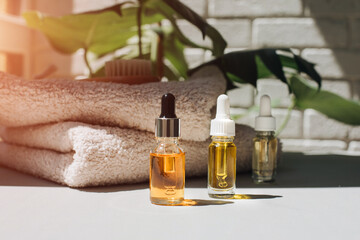 Glass bottles with facial cosmetics liquid on a white table with beige towels and green leaves on backdrop. Front view