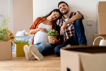 A young happy married couple expecting a baby sitting on the floor of their new home after moving...