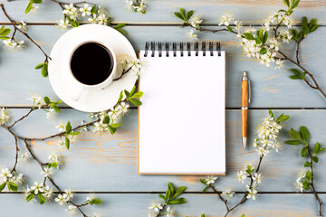 Obraz na płótnie Canvas Artistic composition with opened blank notepad and pen, cup of black coffee, branch of white cherry blossoms on blue wooden table. Festive office desktop concept. Morning coffee cup.