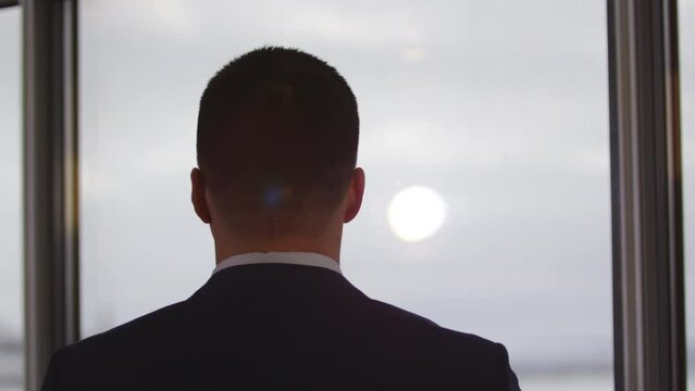 Young businessman stands and watches out of transparent office window back view. Sun illuminates office meeting room brightly