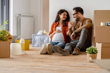 A young happy married couple expecting a baby sitting on the floor of their new home after moving...